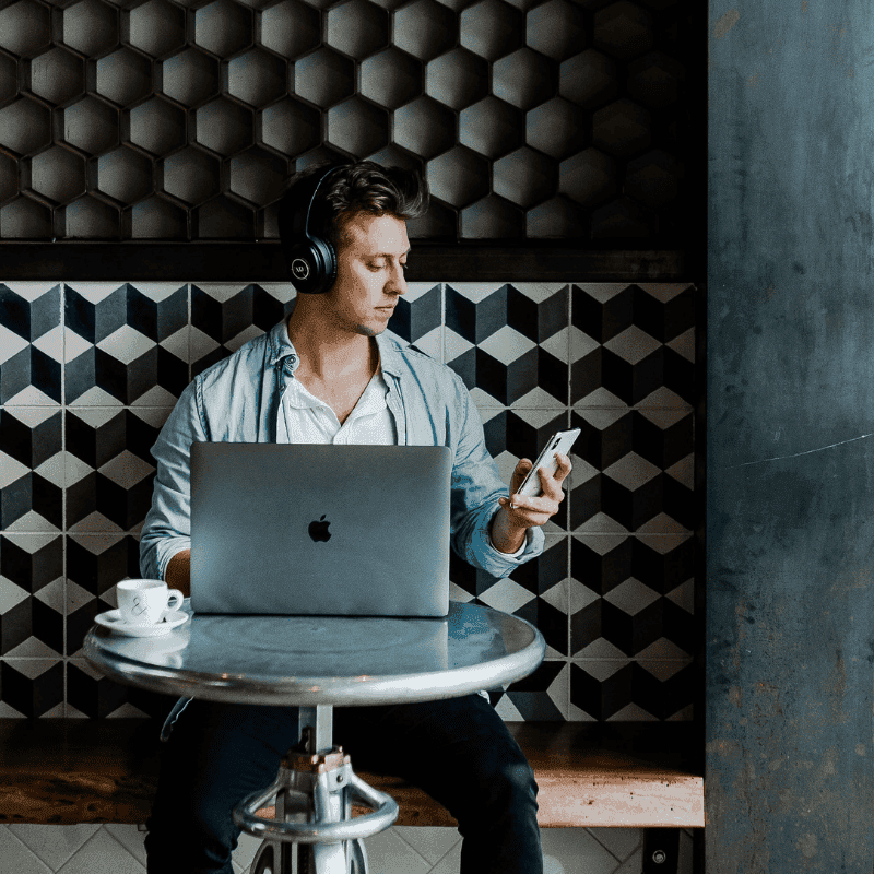 The photo shows a man wearing wireless black headphones, staring down at his white iphone. In front of him on a table is a silver mac with the apple logo showing, and a coffee cup. The scenery is that he could be in a cafe doing some work, researching, or content creating,