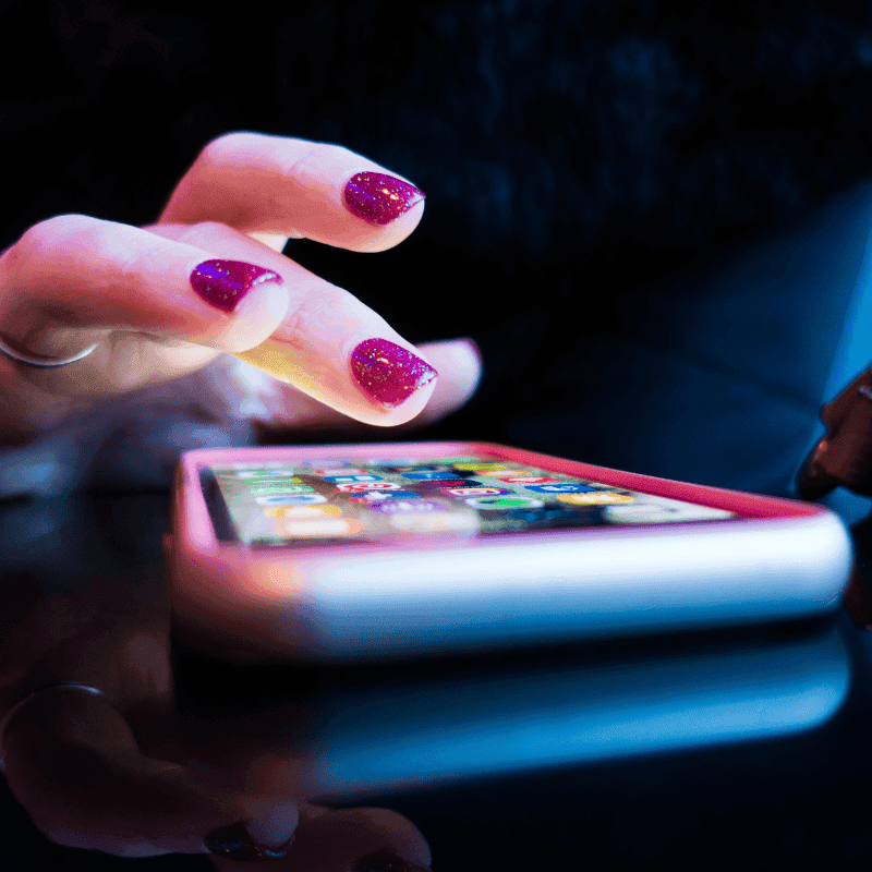 Image shows a very close up photo of a mobile phone on a flat surface. There is a hand above it, looking like it is swiping thriough the phone looking for the correct app. They have pink nail varnish on, covered in glitter, and also a silver shiny ring.