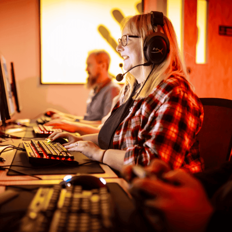 Image shows Jen in a room with red lighting, facing a computer playing a video game. She is wearing a headset with a microphone, like she is playing online with s group of friends. William Cox is in the background, playing on the computer to, but he is slightly faded.