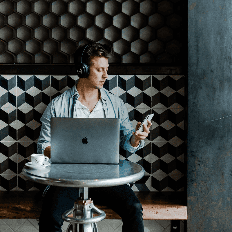 The photo shows a man wearing wireless black headphones, staring down at his white iphone. In front of him on a table is a silver mac with the apple logo showing, and a coffee cup. The scenery is that he could be in a cafe doing some work, researching, or content creating,